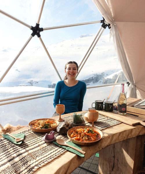 A young white women laughs at a dining table in a clear dome which is her accommodations during a Trolltunga Overnight Hike